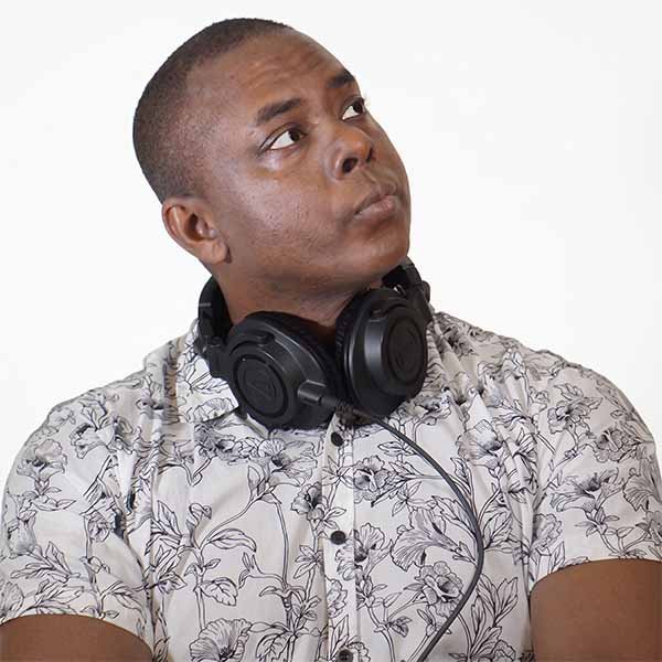 Portrait of DJ Fasmwa wearing headphones around his neck and a patterned floral shirt against a white background