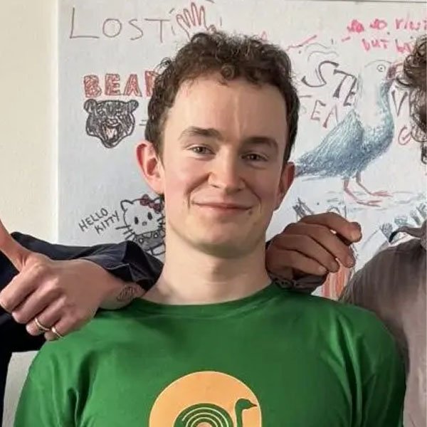 Portrait of Josh Ray, co-host of the 2SER Zine Making Workshop, wearing a green t-shirt in front of an illustrated wall art background.