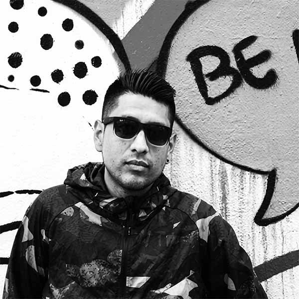 Black-and-white portrait of Paulo Flores (LUNY) wearing sunglasses and a patterned hooded jacket in front of a street art wall.