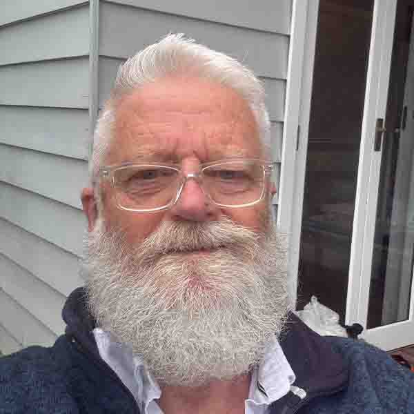 Close-up portrait of Tony Duke of 2SER FM, wearing clear glasses and featuring a full white beard against a light-coloured exterior wall.