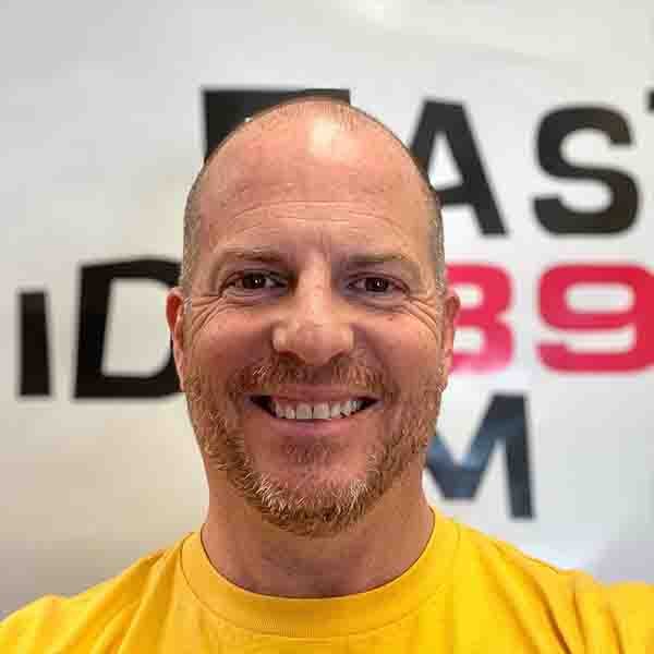 Close-up portrait of Tony Smythe from Eastside Radio smiling in a yellow t-shirt in front of an Eastside 89.7FM station wall logo.