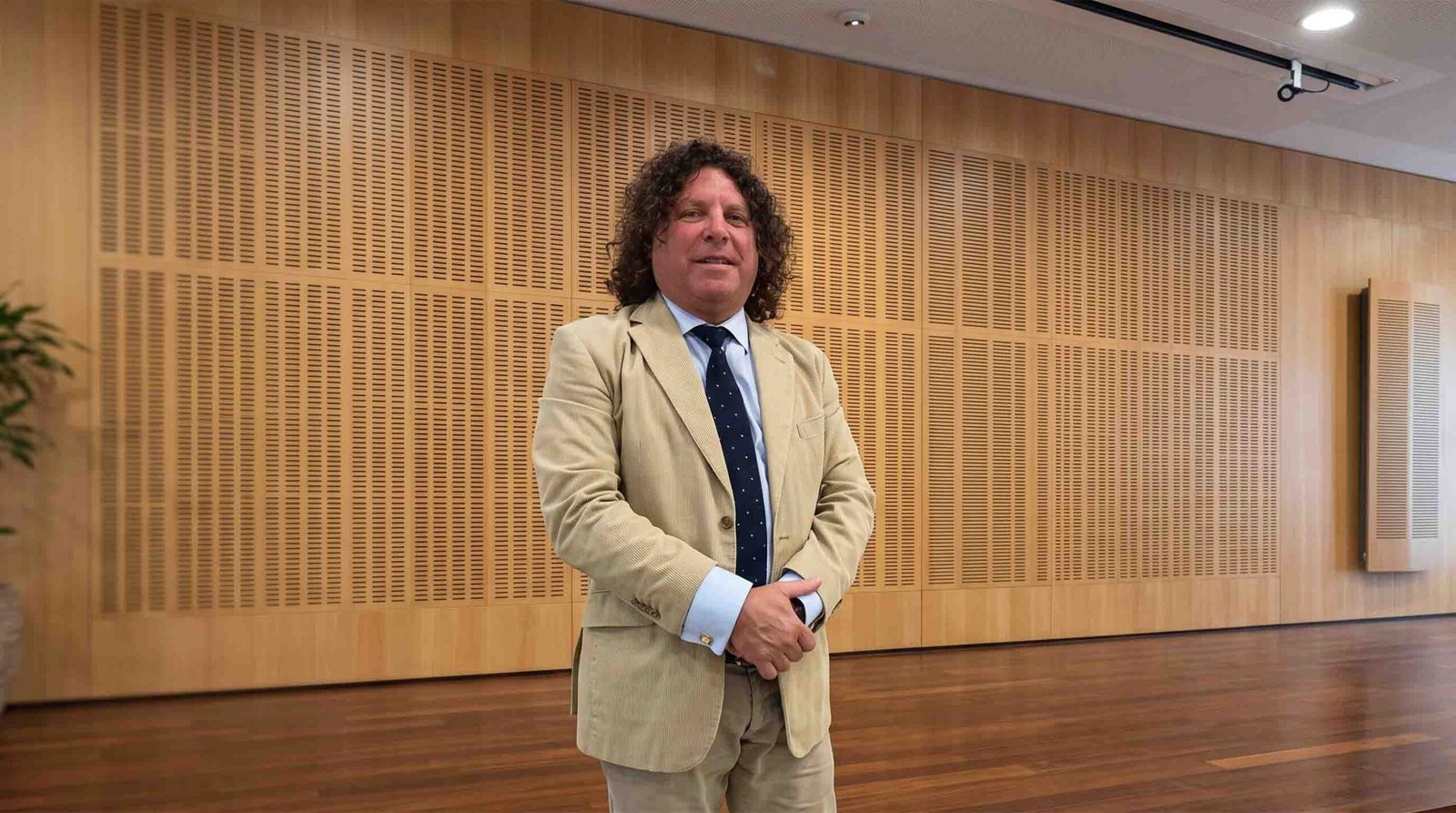 Full-length portrait of Brendan Loizou with dark curly hair, wearing a beige suit jacket, light shirt, and dark blue tie with white polka dots, standing in a modern room with timber-paneled acoustic walls and wooden floors.