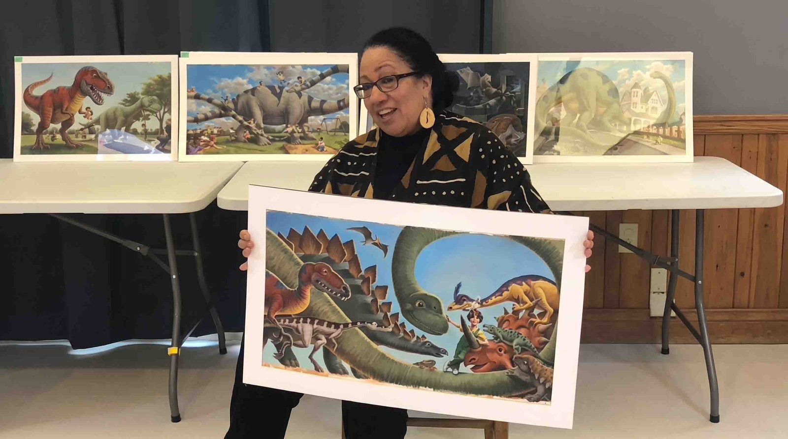 Storyteller Ilene Evans holding a colorful dinosaur illustration print in front of a display of artwork during a workshop.