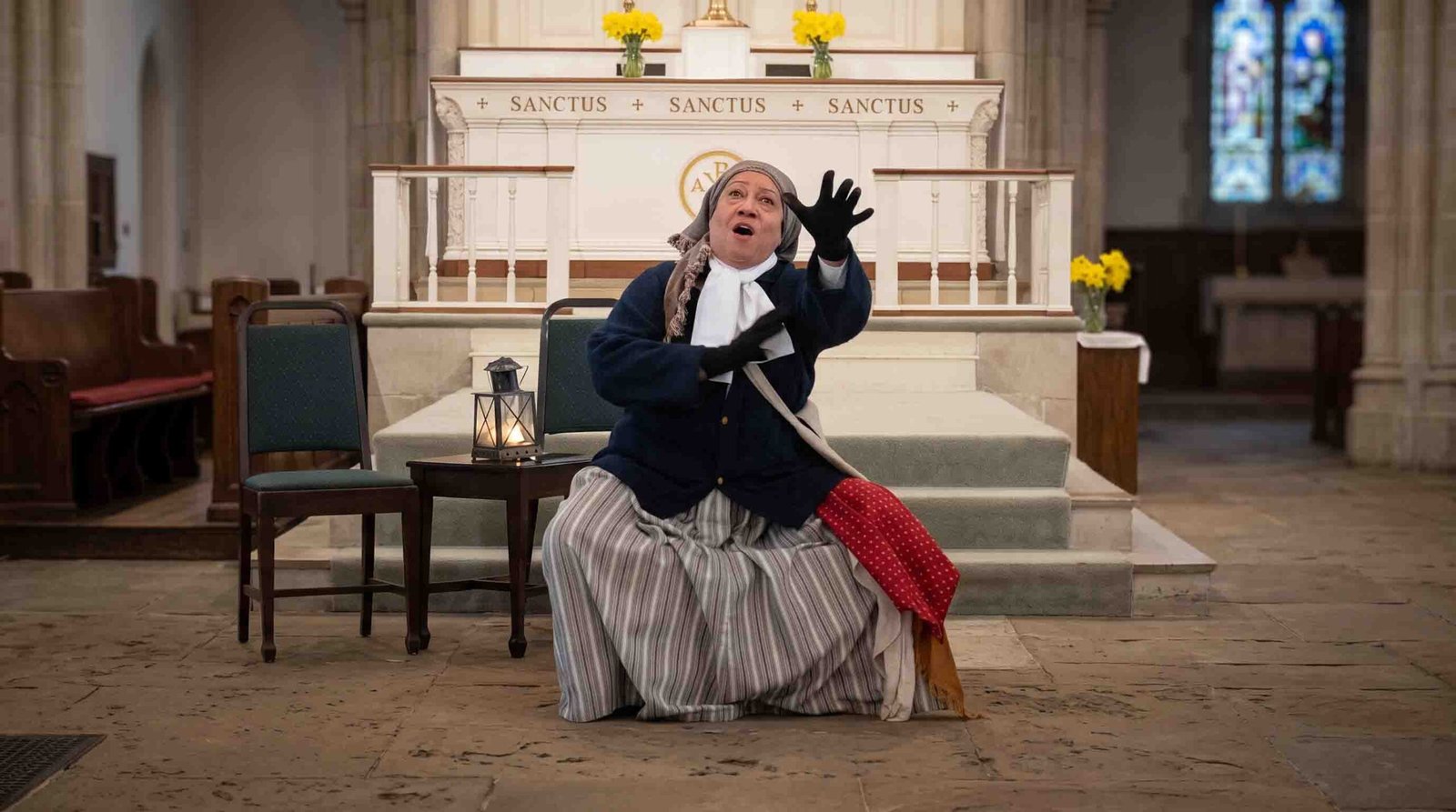 Storyteller Ilene Evans performing in period costume with arm outstretched inside a church sanctuary in front of the altar.