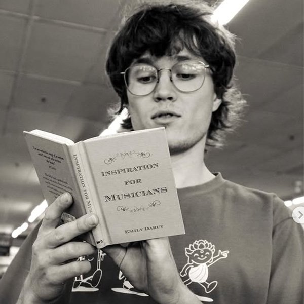 Black-and-white portrait of musician Jude Pascal wearing glasses and reading a book titled "Inspiration for Musicians" by Emily Darcy