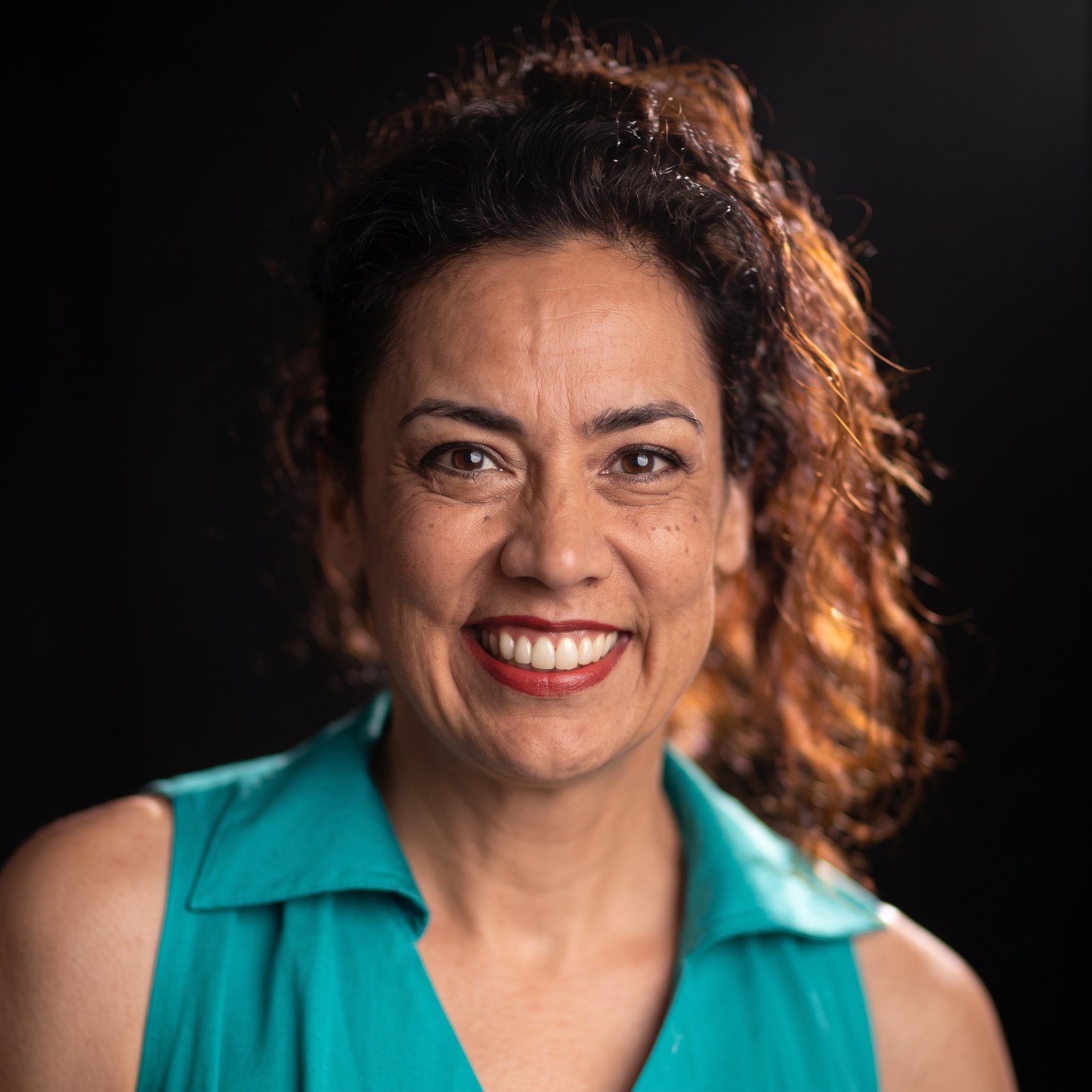 Studio headshot portrait of radio presenter Paula Kruger wearing a teal top and smiling against a dark background.
