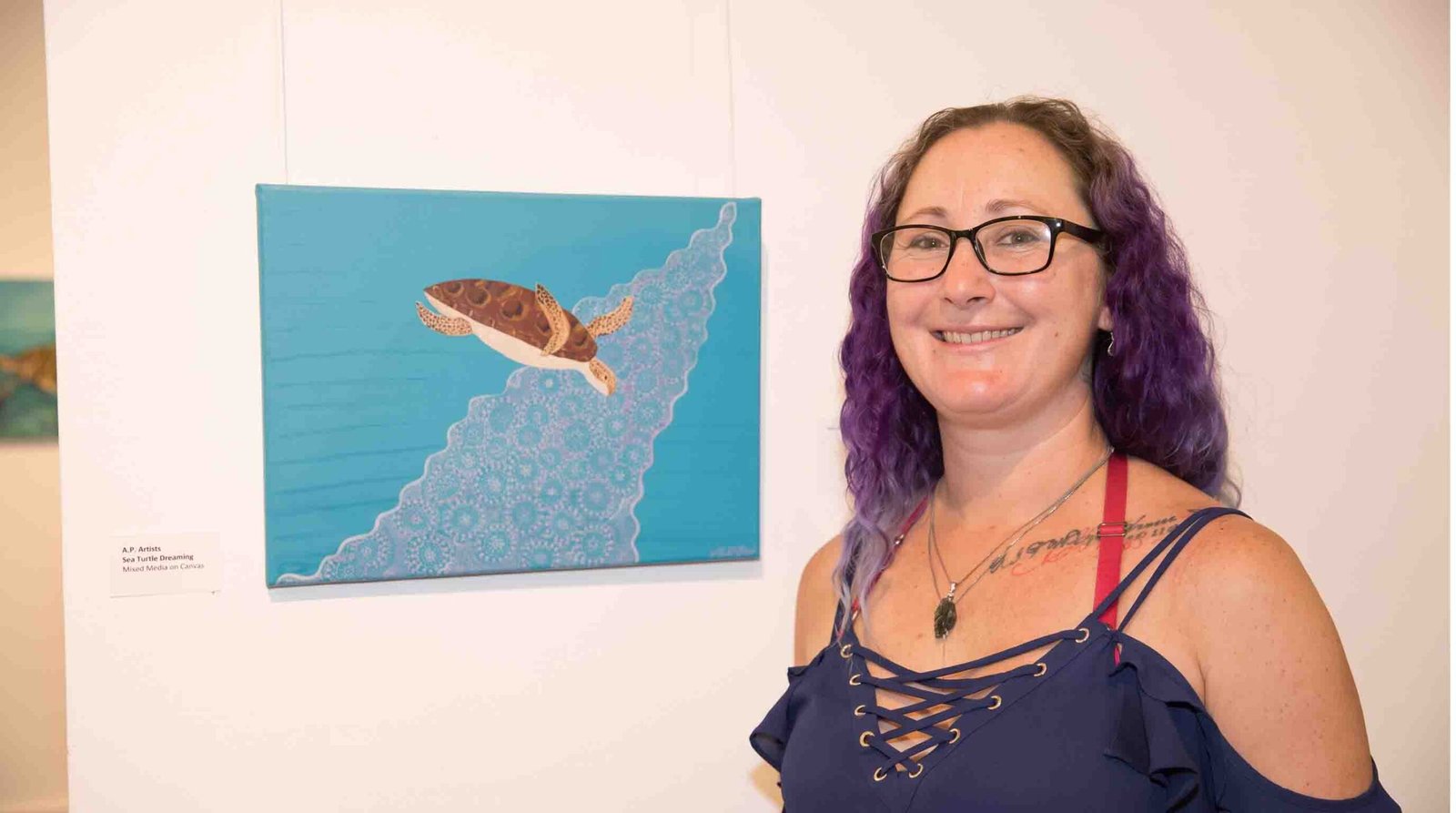 A photograph in UTS Building 1's Main Foyer area of Wiradjuri artist Peta-Joy Williams, with distinct purple hair, smiling broadly as she stands next to her hanging artwork, a canvas featuring a brown sea turtle swimming over a dot-painted wave on a teal background, illustrating 'Sea Turtle Dreaming'.