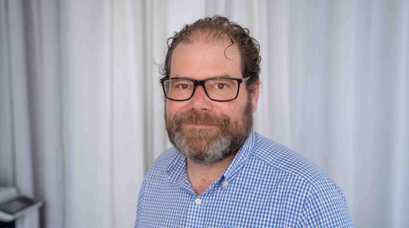 Headshot portrait of Piers Grove wearing glasses and a blue and white checkered button-down shirt, standing in front of light curtains.