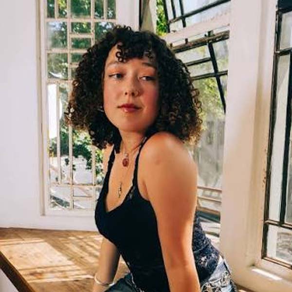 Portrait of musician Ruby Jackson with curly hair, wearing a black lace top and necklace, sitting by a sunny window
